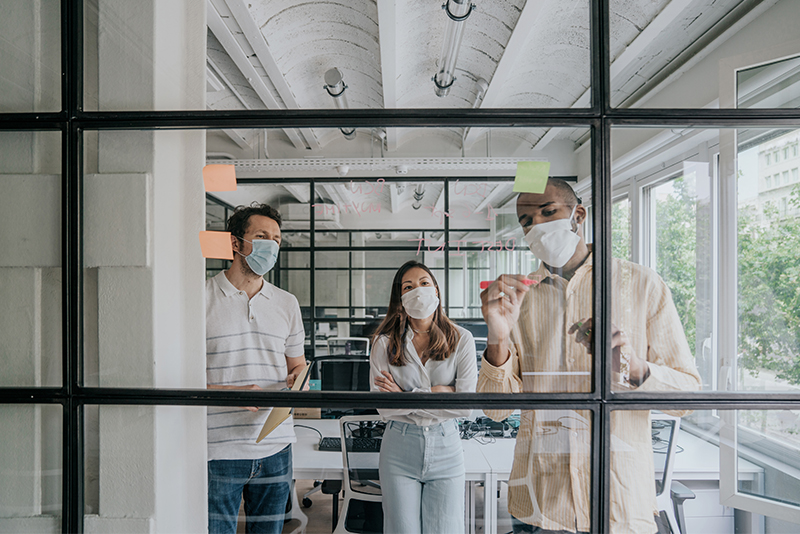 Three adults in casual dress and face masks are writing and collaborating on a business strategy on a interior glass wall in a modern office.