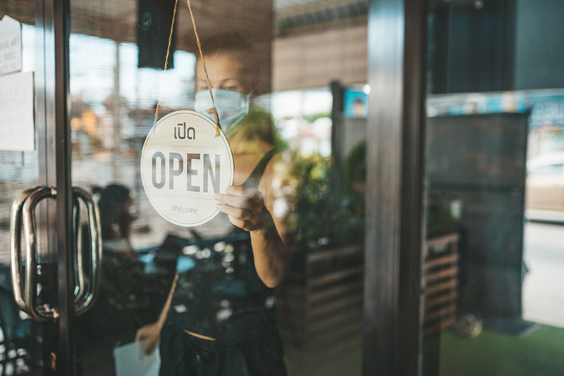 An adult woman in a face mask is flipping the open sign at her retail storefront