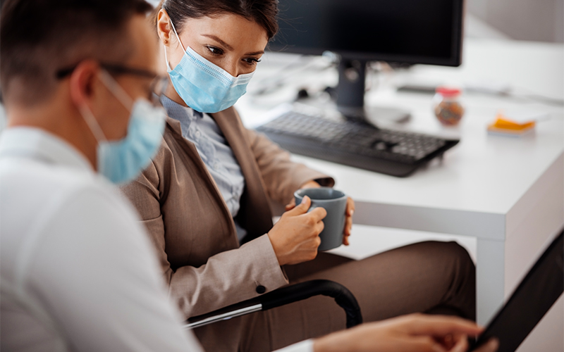 Man and woman in business wear and surgical masks, seated at an office work station, looking at a file together.