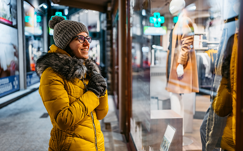 Young woman dressed for winter is window shopping, smiling at something she sees in a shop window.
